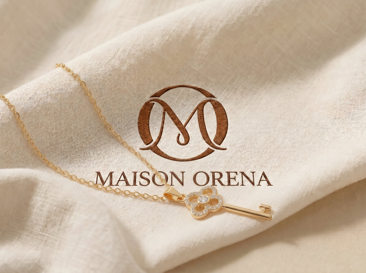 Collier clé trèfle doré posé sur un lin clair, logo Maison Orena en relief